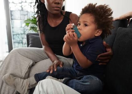 Photo of black child using an asthma inhaler on a couch while the mother supervises